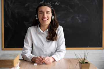 portrait of a happy teacher or tutor sitting in the classroom at the table teaches a lesson online in headphones, headset. Chalk board in the background. Online lecture for college students.