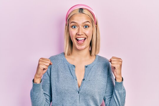 Beautiful blonde woman standing over pink background celebrating surprised and amazed for success with arms raised and open eyes. winner concept.