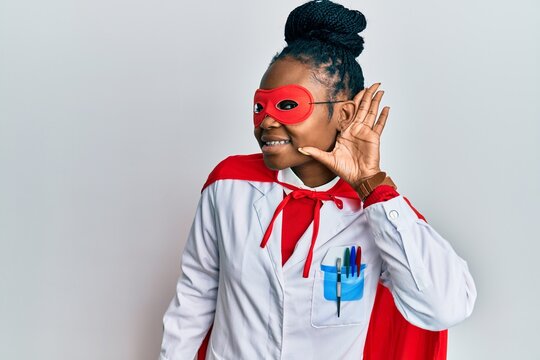 Young african american woman wearing doctor uniform and super hero costume smiling with hand over ear listening and hearing to rumor or gossip. deafness concept.