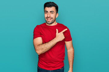 Hispanic man with beard wearing casual red t shirt cheerful with a smile of face pointing with hand and finger up to the side with happy and natural expression on face