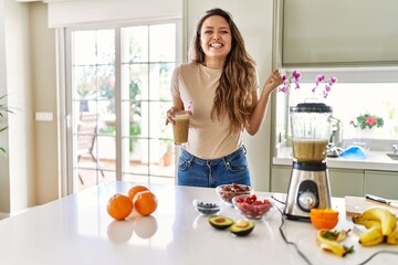 Beautiful young brunette woman drinking glass of smoothie at the kitchen screaming proud,...