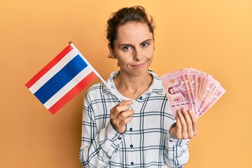 Young brunette woman holding thailand flag and baht banknotes skeptic and nervous, frowning upset because of problem. negative person.