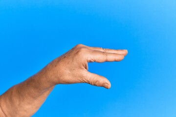Hand of senior hispanic man over blue isolated background picking and taking invisible thing, holding object with fingers showing space