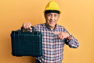 Handsome senior man with grey hair wearing safety helmet holding toolbox smiling happy pointing with hand and finger