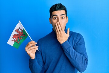 Handsome hispanic man holding wales flag covering mouth with hand, shocked and afraid for mistake....