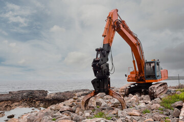 Red excavator with rock or stone grab attached to the arm. Heavy machinery equipment on a construction site by the ocean