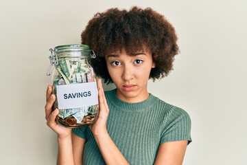Young hispanic girl holding jar with savings clueless and confused expression. doubt concept.