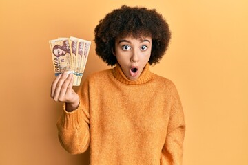 Young hispanic girl holding 5000 hungarian forint banknotes scared and amazed with open mouth for...