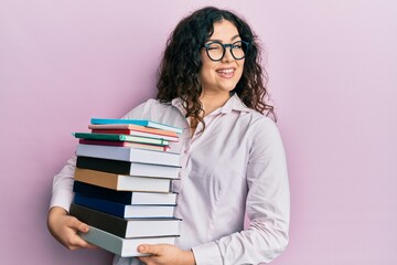 Young brunette woman with curly hair holding a pile of books winking looking at the camera with sexy expression, cheerful and happy face.