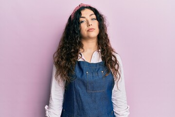 Young brunette woman with curly hair wearing cleaner apron and gloves relaxed with serious expression on face. simple and natural looking at the camera.
