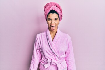Young hispanic woman wearing shower towel cap and bathrobe sticking tongue out happy with funny...
