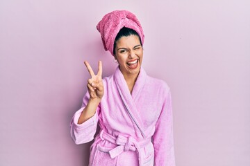 Young hispanic woman wearing shower towel cap and bathrobe smiling with happy face winking at the camera doing victory sign. number two. - obrazy, fototapety, plakaty