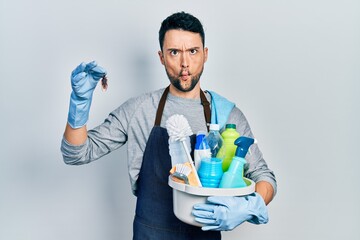 Young hispanic man holding cleaning products and cockroach making fish face with mouth and squinting eyes, crazy and comical.