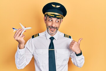 Handsome middle age man with grey hair wearing airplane pilot uniform holding toy plane pointing...