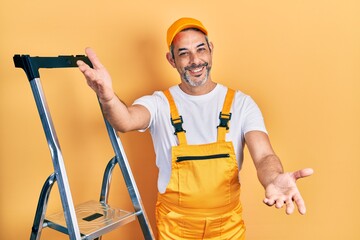 Handsome middle age man with grey hair holding ladder looking at the camera smiling with open arms for hug. cheerful expression embracing happiness.