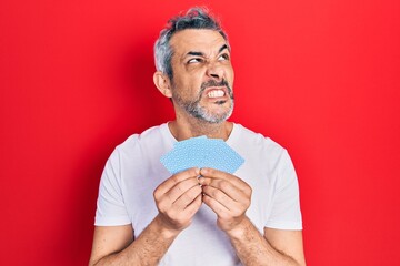 Handsome middle age man with grey hair covering face with cards angry and mad screaming frustrated...