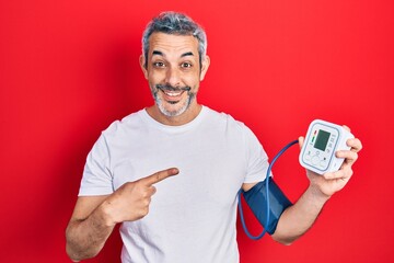 Handsome middle age man with grey hair using blood pressure monitor smiling happy pointing with...