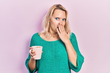 Beautiful caucasian blonde woman drinking a cup of coffee covering mouth with hand, shocked and...