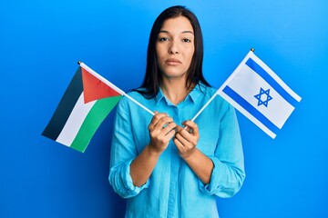 Young latin woman holding palestine and israel flags relaxed with serious expression on face....