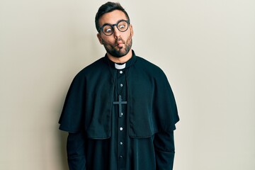 Obraz premium Young hispanic man wearing priest uniform standing over white background making fish face with lips, crazy and comical gesture. funny expression.
