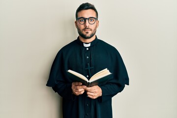 Young hispanic priest man holding bible relaxed with serious expression on face. simple and natural...