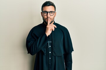 Young hispanic man wearing priest uniform standing over white background asking to be quiet with finger on lips. silence and secret concept.