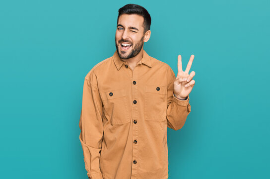 Young hispanic man wearing casual clothes smiling with happy face winking at the camera doing victory sign. number two.