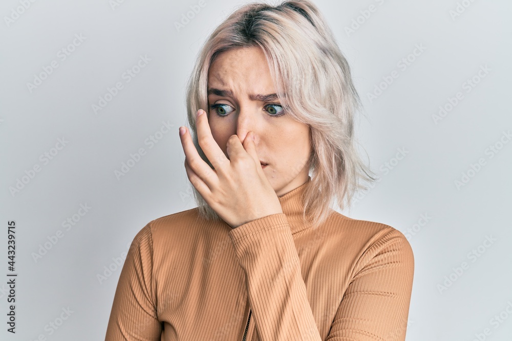 Poster Young blonde girl wearing casual clothes smelling something stinky and disgusting, intolerable smell, holding breath with fingers on nose. bad smell