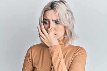 Young blonde girl wearing casual clothes smelling something stinky and disgusting, intolerable smell, holding breath with fingers on nose. bad smell