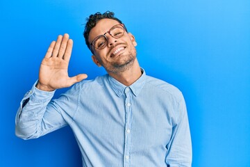 Young hispanic man wearing casual clothes and glasses waiving saying hello happy and smiling,...