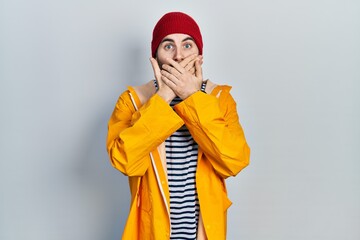 Caucasian man with beard wearing yellow raincoat shocked covering mouth with hands for mistake. secret concept.