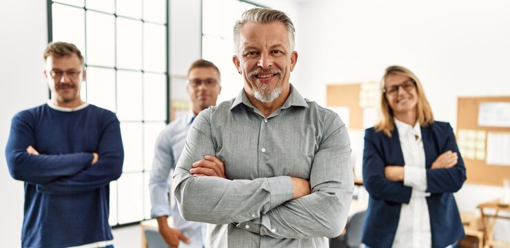 Middle age businessman smiling with arms crossed gesture standing with work partners at the office.