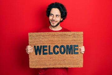 Handsome hispanic man holding welcome doormat smiling looking to the side and staring away thinking.