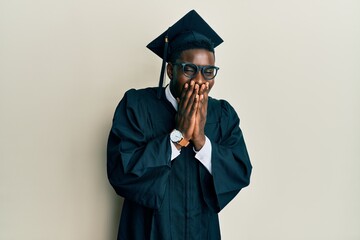 Handsome black man wearing graduation cap and ceremony robe laughing and embarrassed giggle...