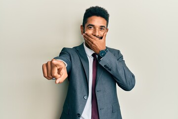 Handsome hispanic man with beard wearing business suit and tie laughing at you, pointing finger to the camera with hand over mouth, shame expression
