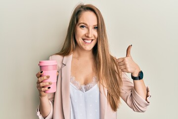 Young blonde woman drinking a take away cup of coffee smiling happy and positive, thumb up doing excellent and approval sign