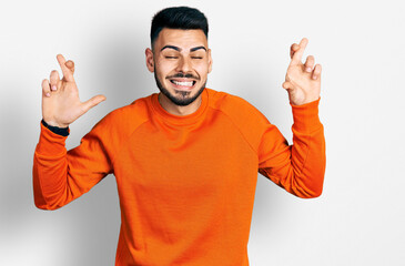 Young hispanic man with beard wearing casual orange sweater gesturing finger crossed smiling with hope and eyes closed. luck and superstitious concept.