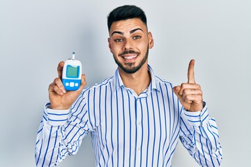 Young hispanic man with beard holding glucometer device smiling with an idea or question pointing...