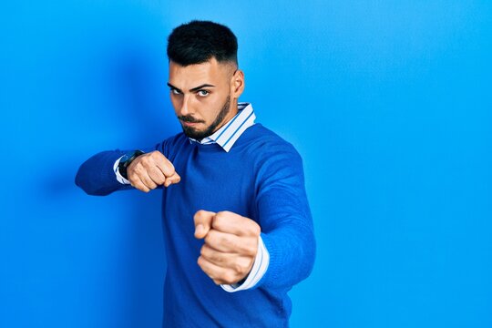 Young hispanic man with beard wearing casual blue sweater punching fist to fight, aggressive and angry attack, threat and violence