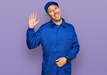 Bald man with beard wearing builder jumpsuit uniform waiving saying hello happy and smiling, friendly welcome gesture