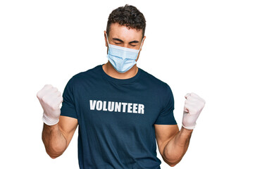 Hispanic young man wearing medical mask and volunteer t shirt very happy and excited doing winner...