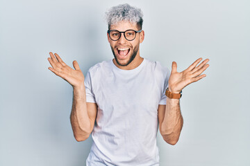 Naklejka premium Young hispanic man with modern dyed hair wearing white t shirt and glasses celebrating crazy and amazed for success with arms raised and open eyes screaming excited. winner concept