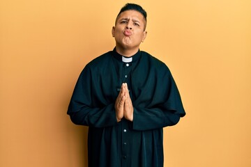 Young latin priest man praying with hands together looking at the camera blowing a kiss being...