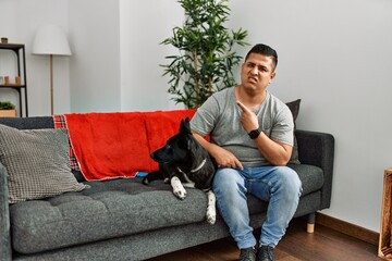 Young latin man and dog sitting on the sofa at home pointing aside worried and nervous with forefinger, concerned and surprised expression