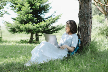 Nice teenage girl siting near tree in summer park with open laptop in her hands and studying. Concept of distance learning