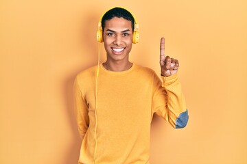 Young african american guy listening to music using headphones showing and pointing up with finger number one while smiling confident and happy.