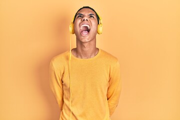 Obraz premium Young african american guy listening to music using headphones angry and mad screaming frustrated and furious, shouting with anger. rage and aggressive concept.
