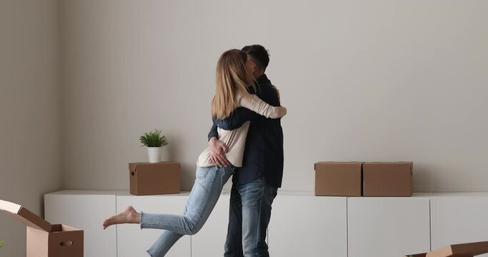 Happy millennial 30s married couple celebrate relocation day to new own house, bank loan and mortgage, marriage concept. Loving husband lifts wife, homeowner family cuddling swirling looking overjoyed