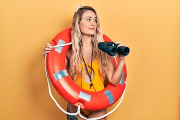 Beautiful young blonde woman wearing bikini and holding lifeguard float and binoculars smiling looking to the side and staring away thinking.