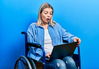 Beautiful caucasian woman sitting on wheelchair using laptop scared and amazed with open mouth for...